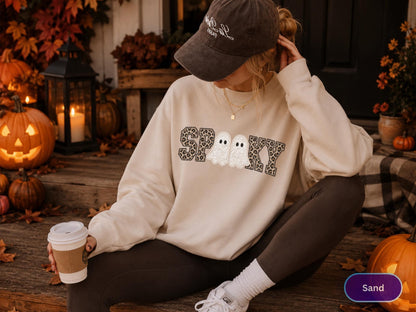 May include: A beige crewneck sweatshirt featuring the word SPOOKY in a black and grey leopard print pattern, with two white ghost illustrations replacing the letter O. The garment has long sleeves and a relaxed fit, worn with dark leggings.