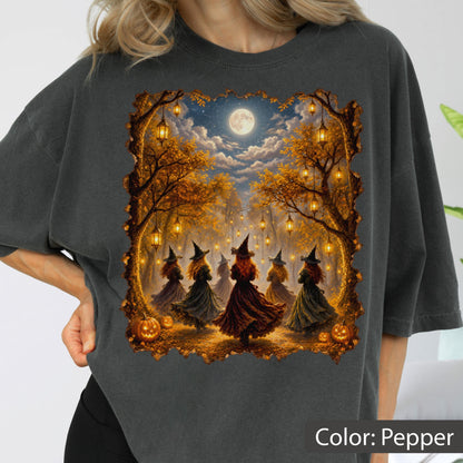 May include: A charcoal gray t-shirt featuring a graphic print of five figures in witch hats and long dresses standing in a forest clearing under a full moon. The scene includes hanging lanterns, autumn trees, and pumpkins on the ground.