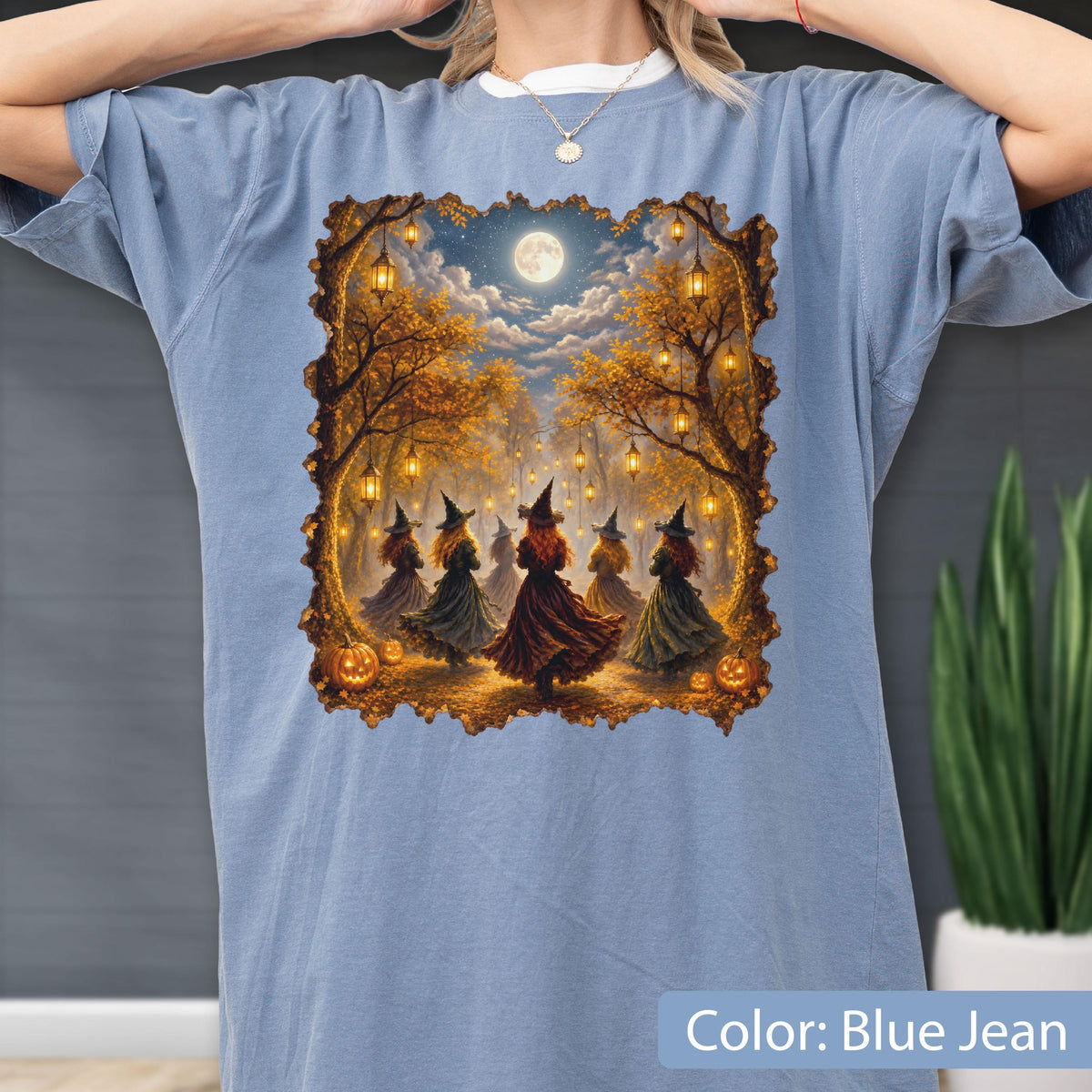 May include: A blue jean colored t-shirt featuring a rectangular graphic print of five figures wearing pointed hats and long dresses standing in an autumn forest at night. The scene includes hanging lanterns, a full moon, and orange pumpkins.
