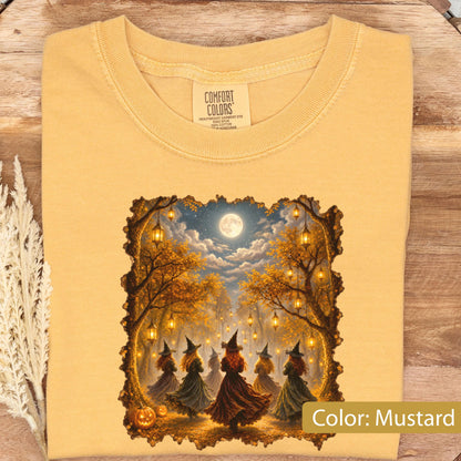 May include: A mustard yellow t-shirt with a Comfort Colors label at the neck. The graphic features a group of witches in long dresses standing in a forest at night under a full moon, surrounded by hanging lanterns and autumn trees with orange leaves.