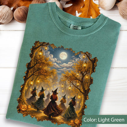 May include: A light green crew neck t-shirt featuring a graphic print of several figures in pointed hats dancing in a forest at night under a full moon. The scene includes glowing lanterns hanging from autumn trees and orange pumpkins on the ground.
