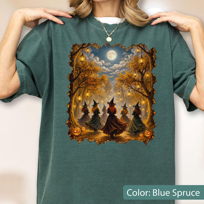 May include: A dark teal short-sleeve t-shirt featuring a rectangular graphic print of five figures wearing pointed hats and long dresses dancing in a forest under a full moon. The scene includes hanging lanterns, autumn trees, and orange pumpkins.