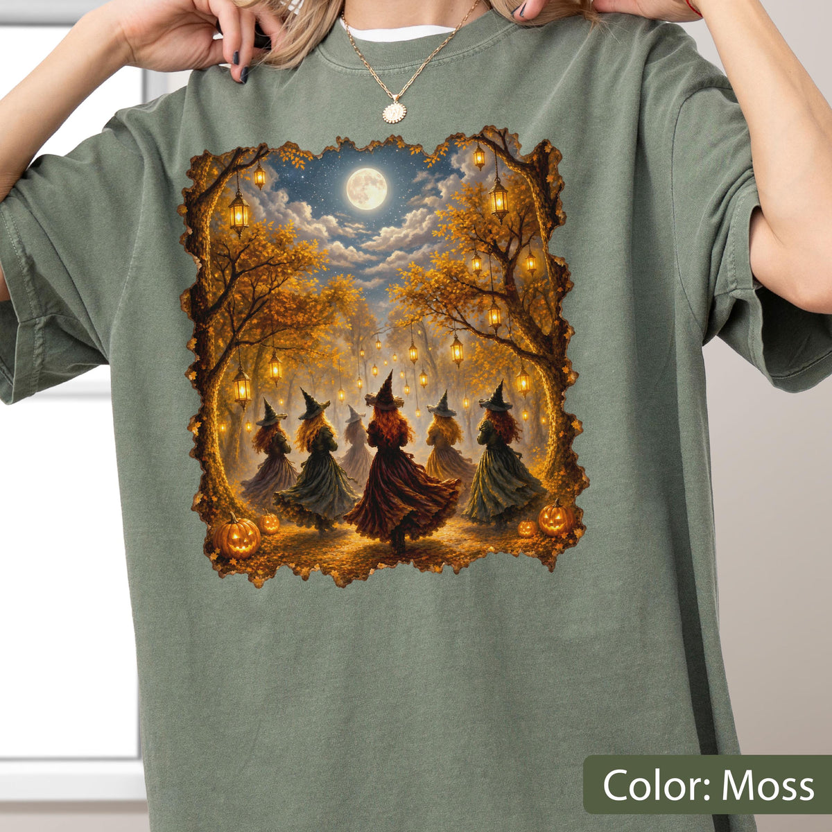 May include: A moss green short-sleeve t-shirt featuring a rectangular graphic print of five figures wearing pointed hats and long dresses dancing in a forest at night. The scene includes glowing lanterns hanging from trees, orange pumpkins, and a full moon.