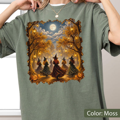 May include: A moss green short-sleeve t-shirt featuring a rectangular graphic print of five figures wearing pointed hats and long dresses dancing in a forest at night. The scene includes glowing lanterns hanging from trees, orange pumpkins, and a full moon.