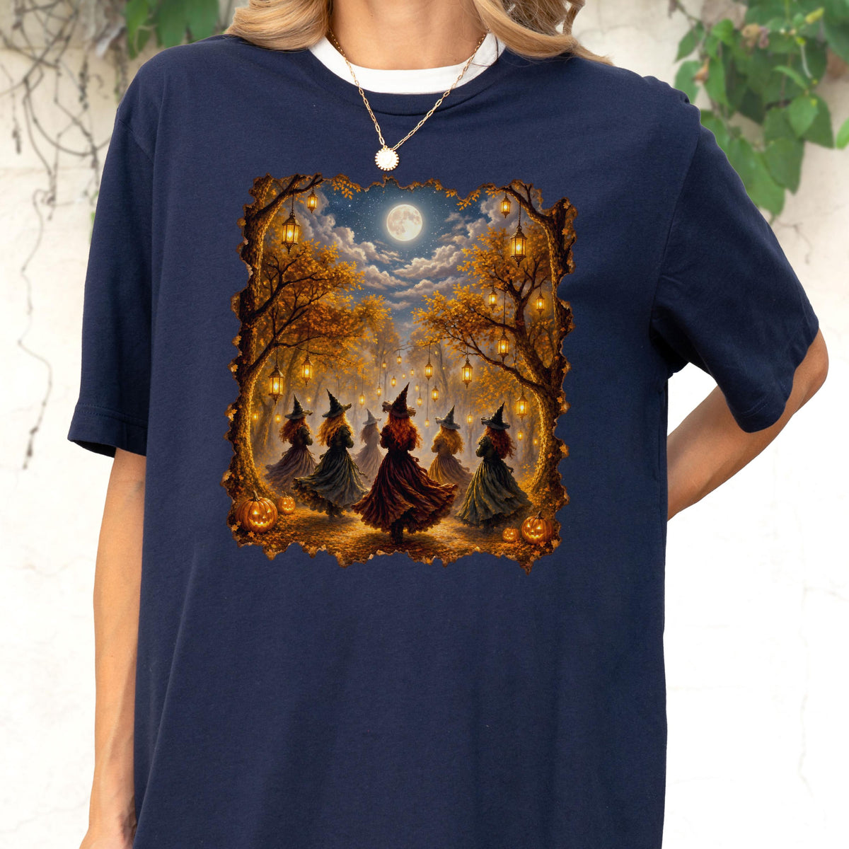 May include: A navy blue t-shirt featuring a graphic print of several figures wearing pointed hats and long dresses standing in a forest at night. The scene includes glowing lanterns hanging from trees, a full moon in the sky, and orange pumpkins.