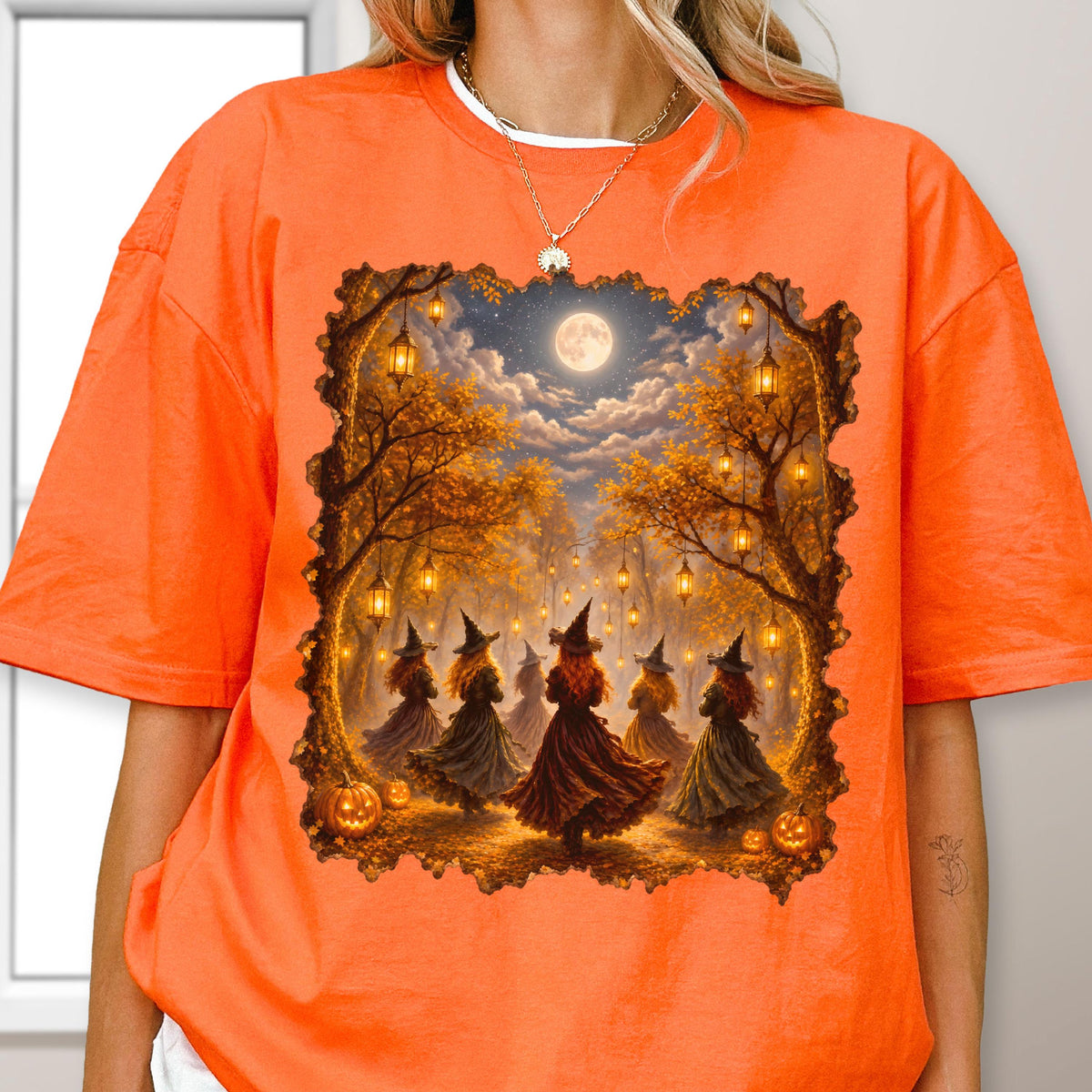 May include: An orange t-shirt featuring a rectangular graphic print of a forest scene at night with a full moon, hanging lanterns, and several figures wearing pointed hats and long dresses, surrounded by autumn trees and pumpkins.