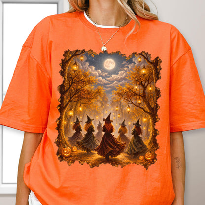 May include: An orange t-shirt featuring a rectangular graphic print of a forest scene at night with a full moon, hanging lanterns, and several figures wearing pointed hats and long dresses, surrounded by autumn trees and pumpkins.