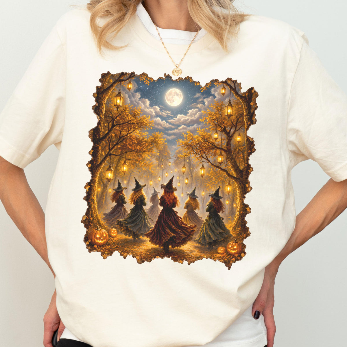May include: A cream-colored short-sleeve t-shirt featuring a rectangular graphic print of several figures in pointed hats dancing in a forest at night. The scene includes glowing lanterns hanging from autumn trees, a full moon, and orange pumpkins.