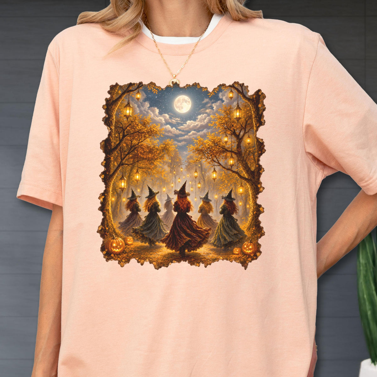 May include: A light peach-colored t-shirt featuring a rectangular graphic print of several figures wearing pointed hats and long dresses dancing in a forest at night. The scene includes glowing lanterns hanging from trees, pumpkins, and a full moon.