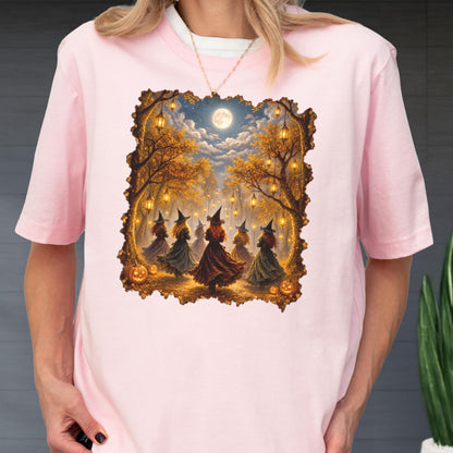May include: A light pink short-sleeved t-shirt featuring a rectangular graphic print of several figures wearing pointed hats and long dresses dancing in a forest at night. The scene includes glowing lanterns hanging from trees, pumpkins, and a full moon.