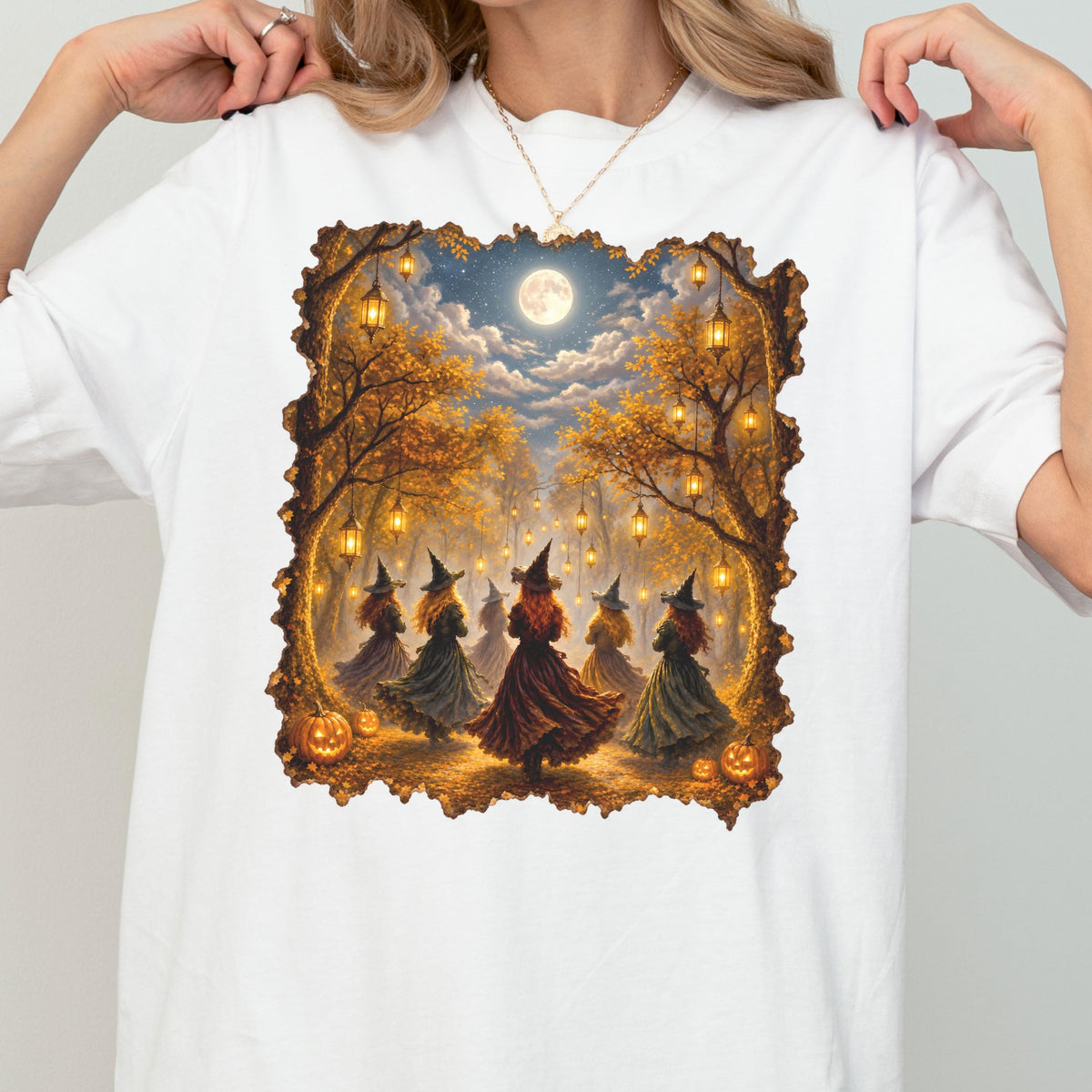 May include: A white t-shirt featuring a rectangular graphic print of a group of figures in pointed hats dancing in an autumn forest at night. The scene includes glowing lanterns hanging from trees, a full moon, and orange pumpkins on the ground.
