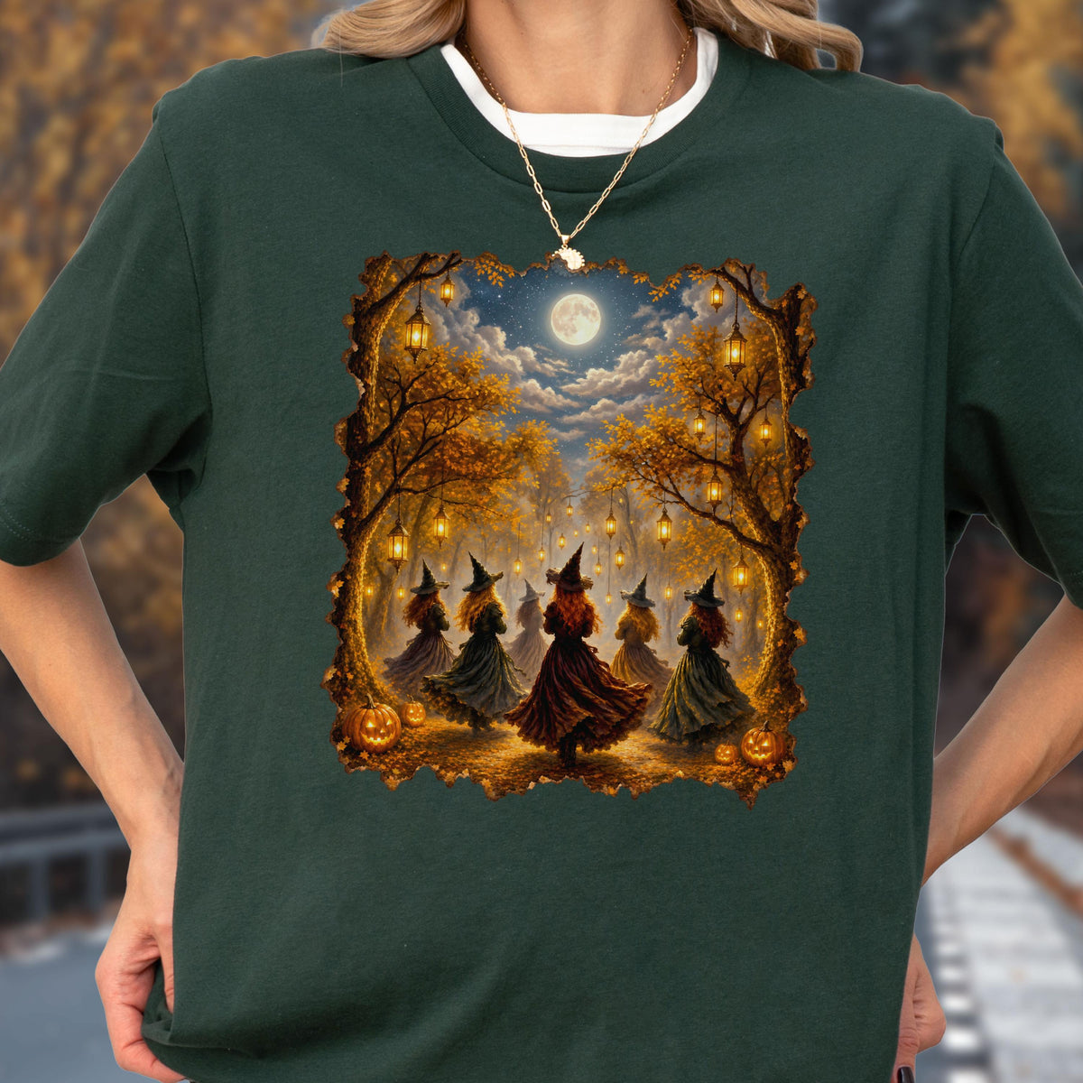 May include: A dark green short-sleeved t-shirt featuring a rectangular graphic print of five figures in pointed hats and long dresses standing in a forest at night under a full moon, surrounded by hanging lanterns and orange pumpkins on the ground.