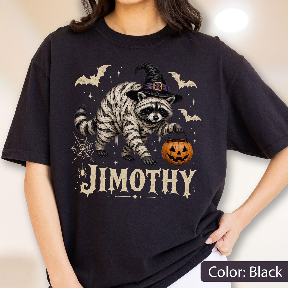 May include: A black t-shirt featuring a graphic of a raccoon wrapped in white bandages, wearing a black witch hat, and holding an orange jack-o'-lantern bucket. The design includes bats, a spider web, and the text JIMOTHY in a large serif font.