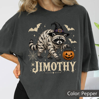 May include: A charcoal gray t-shirt featuring a graphic of a raccoon wrapped in white bandages like a mummy, wearing a black witch hat, and holding an orange jack-o'-lantern bucket. The design includes bats, a spider web, and the text JIMOTHY.