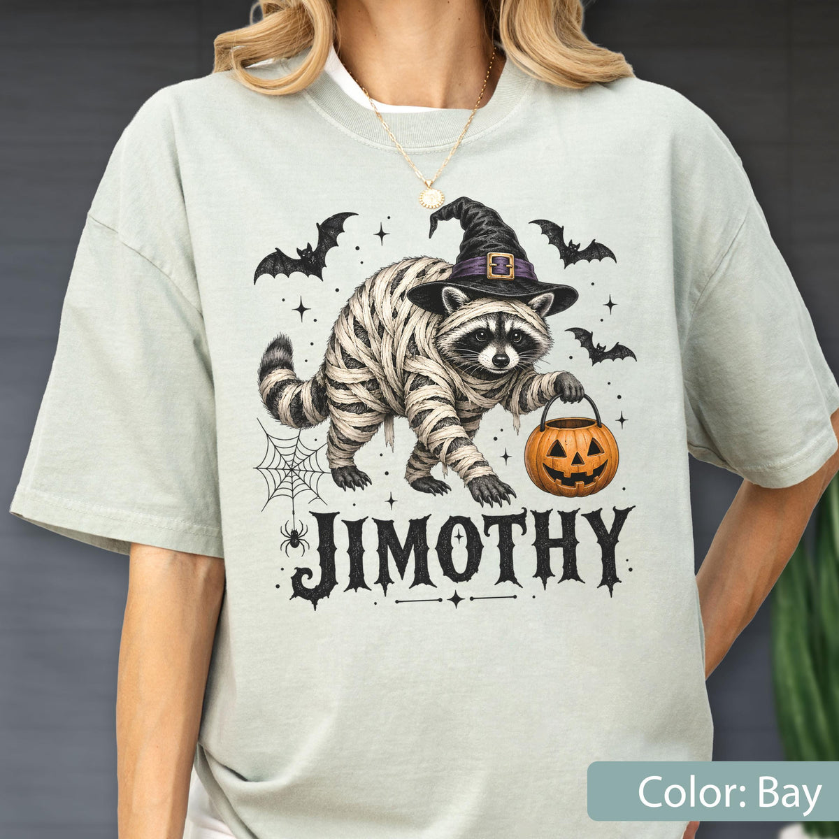 May include: A light sage green t-shirt featuring a graphic of a raccoon wrapped in mummy bandages, wearing a black witch hat, and holding an orange jack-o'-lantern. The design includes bats, a spider web, and the text JIMOTHY in black lettering.