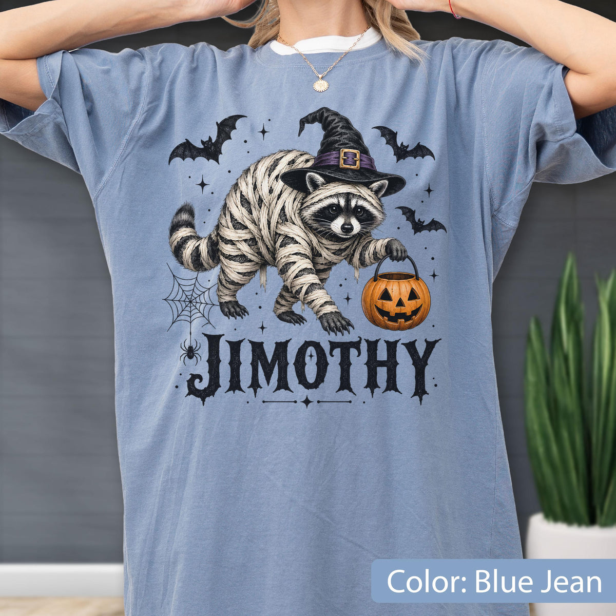 May include: A blue jean colored t-shirt featuring a graphic of a raccoon wrapped in bandages like a mummy, wearing a black witch hat, and holding an orange jack-o'-lantern bucket. The text JIMOTHY is printed below with bats and a spider web.
