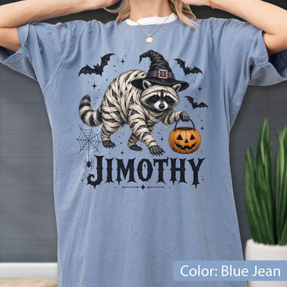 May include: A blue jean colored t-shirt featuring a graphic of a raccoon wrapped in bandages like a mummy, wearing a black witch hat, and holding an orange jack-o'-lantern bucket. The text JIMOTHY is printed below with bats and a spider web.