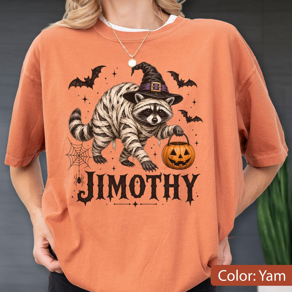 May include: An orange t-shirt featuring a graphic of a raccoon wrapped in bandages like a mummy, wearing a black witch hat, and holding a jack-o'-lantern bucket. The design includes bats, a spider web, and the text JIMOTHY in black lettering.