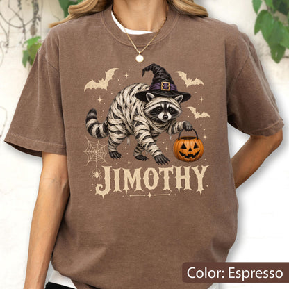 May include: An espresso-colored t-shirt featuring a graphic of a raccoon wrapped in white bandages, wearing a black witch hat, and holding an orange jack-o'-lantern. The design includes bats, a spider web, and the name JIMOTHY in large letters.