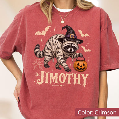 May include: A crimson t-shirt featuring a graphic of a raccoon wrapped in bandages like a mummy, wearing a black witch hat and holding a jack-o'-lantern bucket. The design includes bats, a spider web, and the text JIMOTHY in a decorative font.
