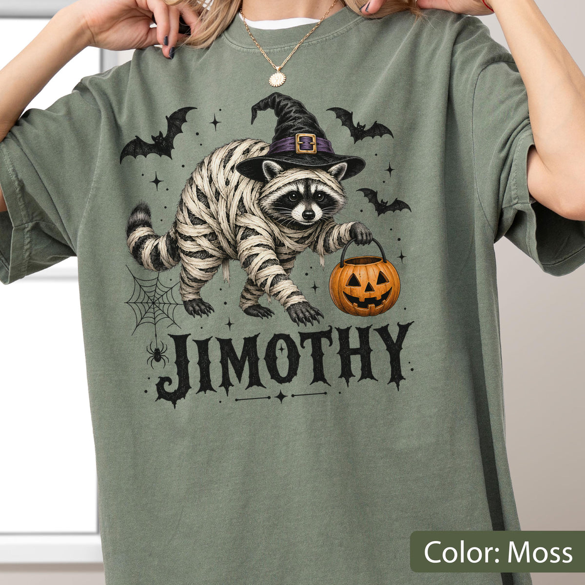 May include: A moss green t-shirt featuring a graphic of a raccoon wrapped in mummy bandages, wearing a black witch hat, and holding an orange jack-o'-lantern. The design includes black bats, a spider web, and the name JIMOTHY in black text.