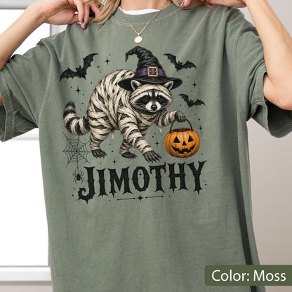 May include: A moss green t-shirt featuring a graphic of a raccoon wrapped in mummy bandages, wearing a black witch hat, and holding an orange jack-o'-lantern. The design includes black bats, a spider web, and the name JIMOTHY in black text.
