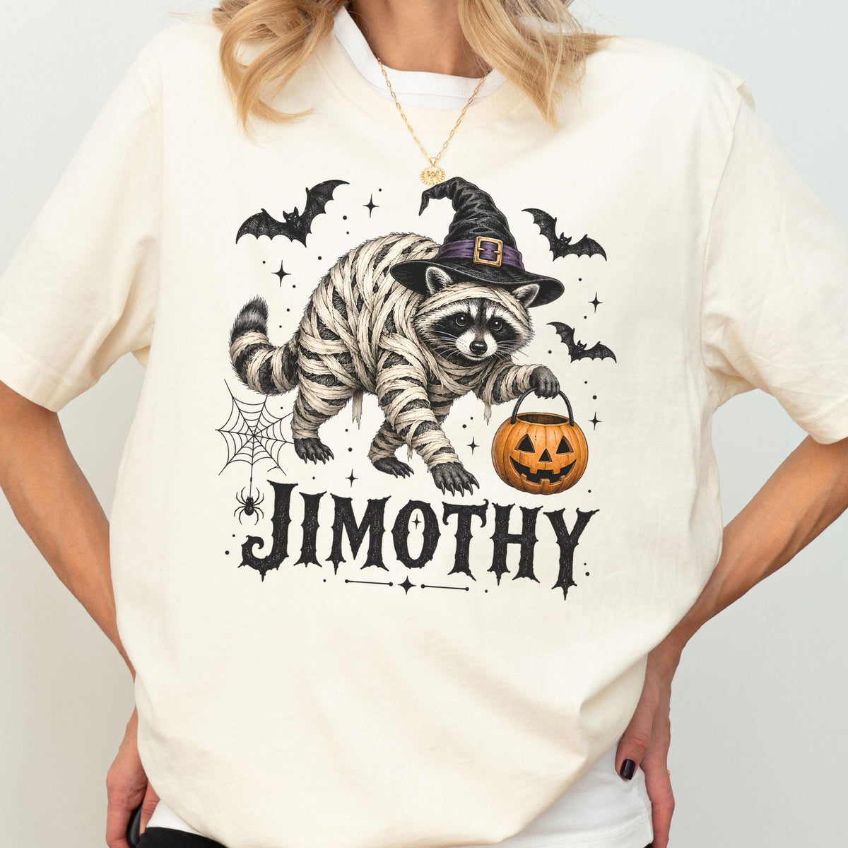 May include: A cream-colored t-shirt featuring a graphic of a raccoon dressed as a mummy, wearing a black witch hat, and holding an orange jack-o'-lantern bucket. The design includes bats, a spider web, and the name JIMOTHY in black lettering.