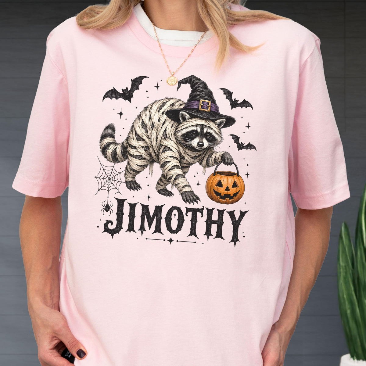May include: A light pink t-shirt featuring a graphic of a raccoon wrapped in mummy bandages, wearing a black witch hat, and holding an orange jack-o'-lantern. The design includes bats, a spider web, and the name JIMOTHY in black lettering.