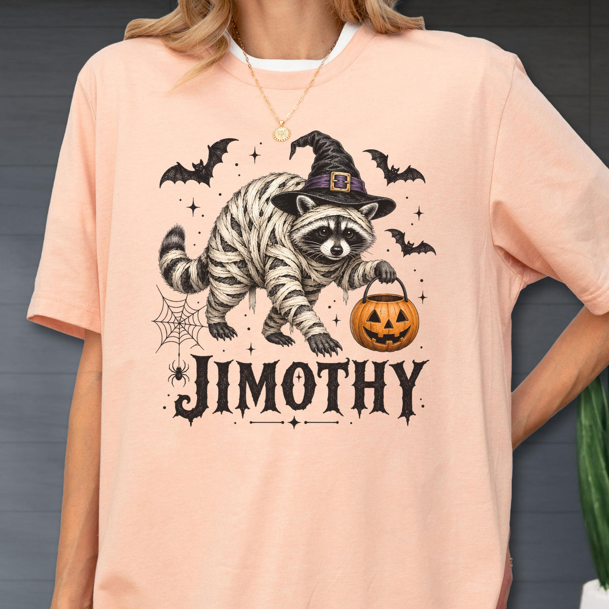 May include: A peach-colored t-shirt featuring a graphic of a raccoon wrapped in mummy bandages, wearing a black witch hat, and holding an orange jack-o'-lantern bucket. The design includes bats, a spider web, and the text JIMOTHY in black.