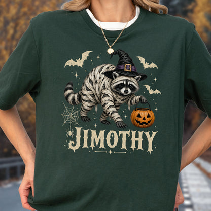 May include: A dark green t-shirt featuring a graphic of a raccoon wrapped in white bandages like a mummy, wearing a black witch hat, and holding an orange jack-o'-lantern bucket. The design includes bats, a spider web, and the text JIMOTHY.