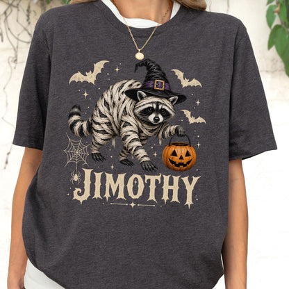 May include: A charcoal gray t-shirt featuring a graphic of a raccoon wrapped in white bandages, wearing a black witch hat, and holding an orange jack-o'-lantern bucket. The design includes bats, a spider web, and the text JIMOTHY in cream.