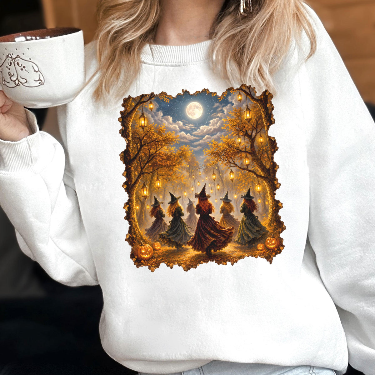 May include: A white crewneck sweatshirt featuring a rectangular graphic print of five figures in pointed hats and long dresses dancing in a forest at night. The scene includes glowing lanterns hanging from trees, a full moon, and orange pumpkins.