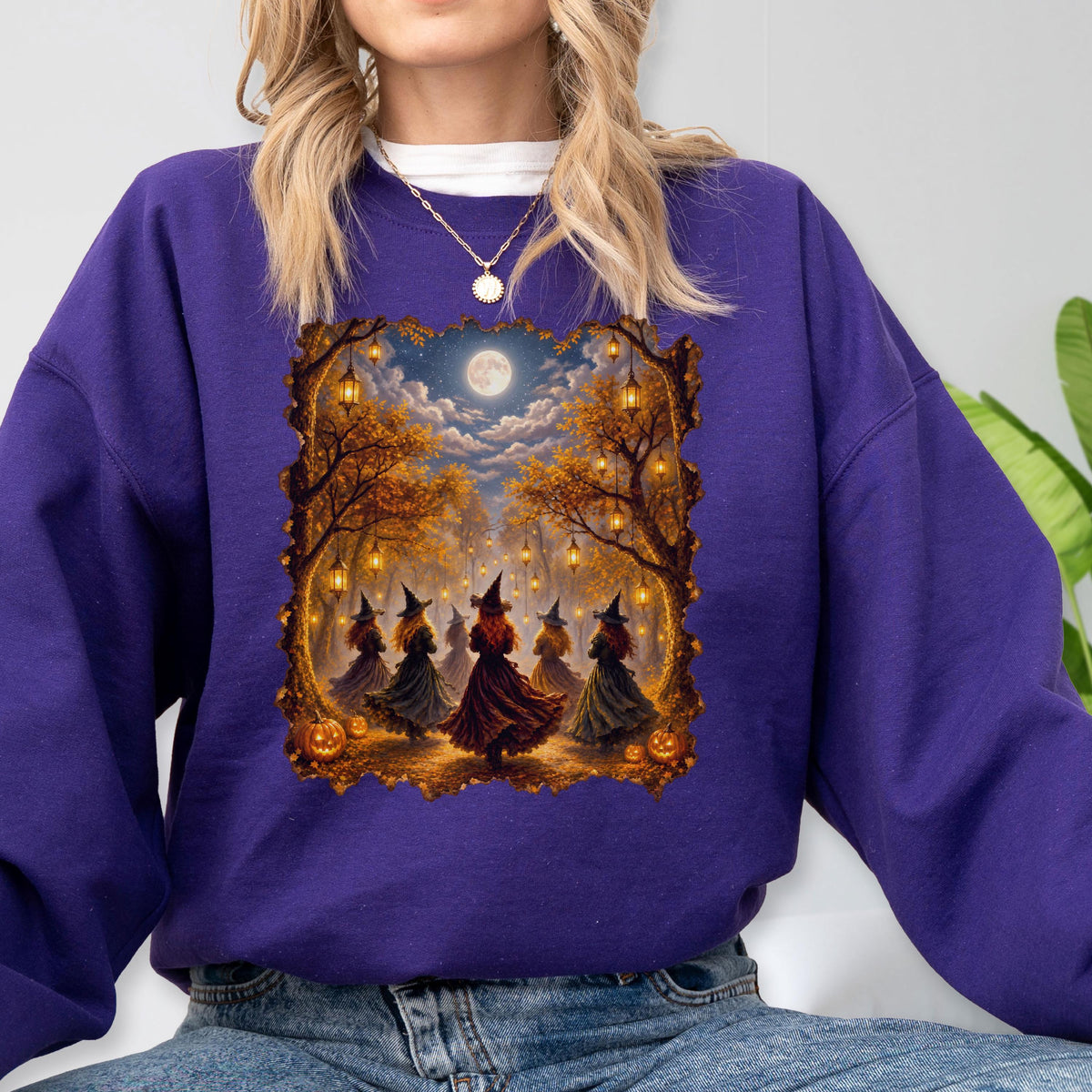May include: A purple long-sleeve sweatshirt featuring a rectangular graphic print of five figures wearing pointed hats and dresses standing in a forest with hanging lanterns, a full moon, and orange pumpkins under a cloudy night sky.
