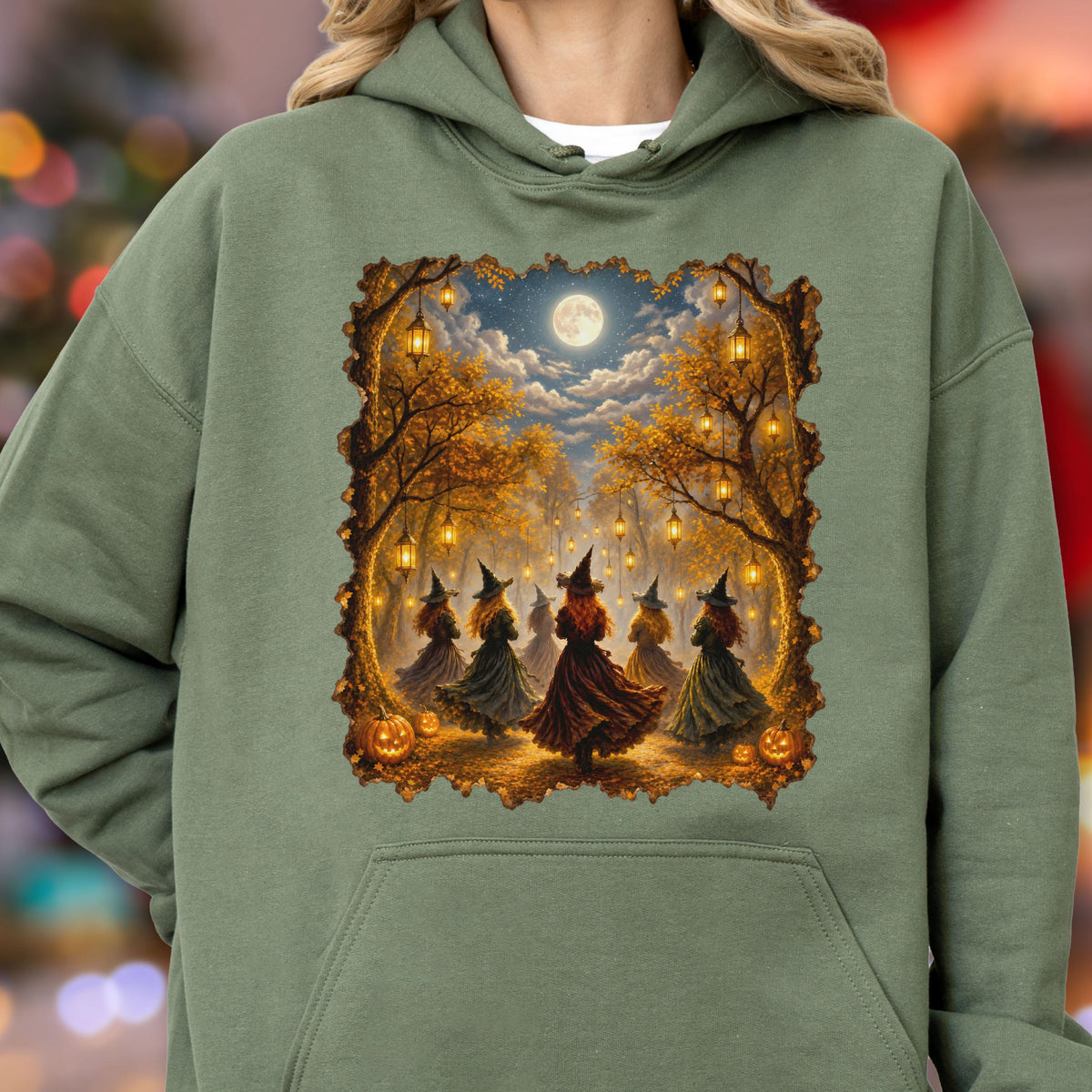 May include: An olive green hooded sweatshirt featuring a rectangular graphic print of five figures wearing pointed hats and long dresses standing in a forest at night. The scene includes hanging lanterns, autumn trees, a full moon, and pumpkins.