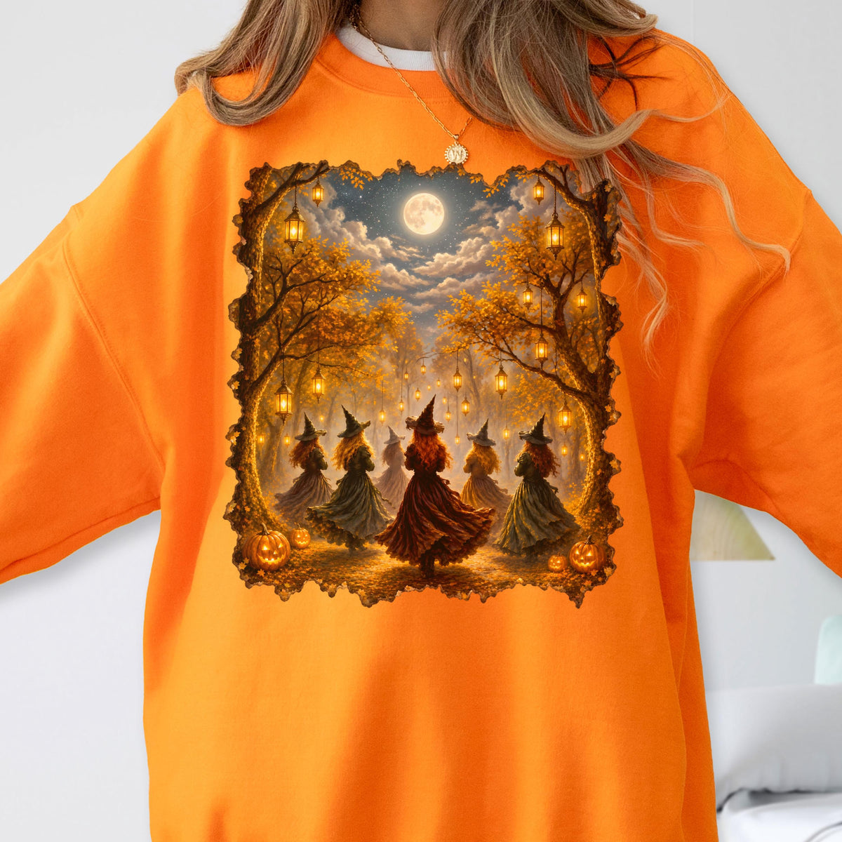 May include: A bright orange sweatshirt featuring a rectangular graphic print of five figures in pointed hats standing in a forest under a full moon. The scene includes hanging lanterns, autumn trees with golden leaves, and pumpkins on the ground.