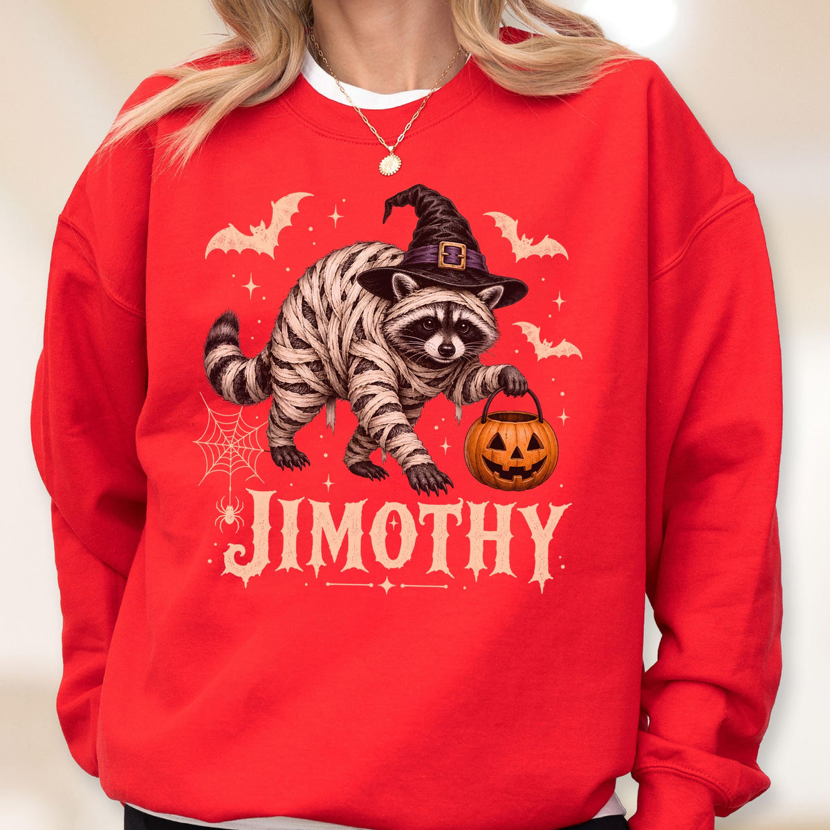 May include: A bright red crewneck sweatshirt featuring a graphic of a raccoon wrapped in mummy bandages, wearing a black witch hat, and holding an orange jack-o'-lantern bucket. The design includes bats, a spider web, and the text JIMOTHY.