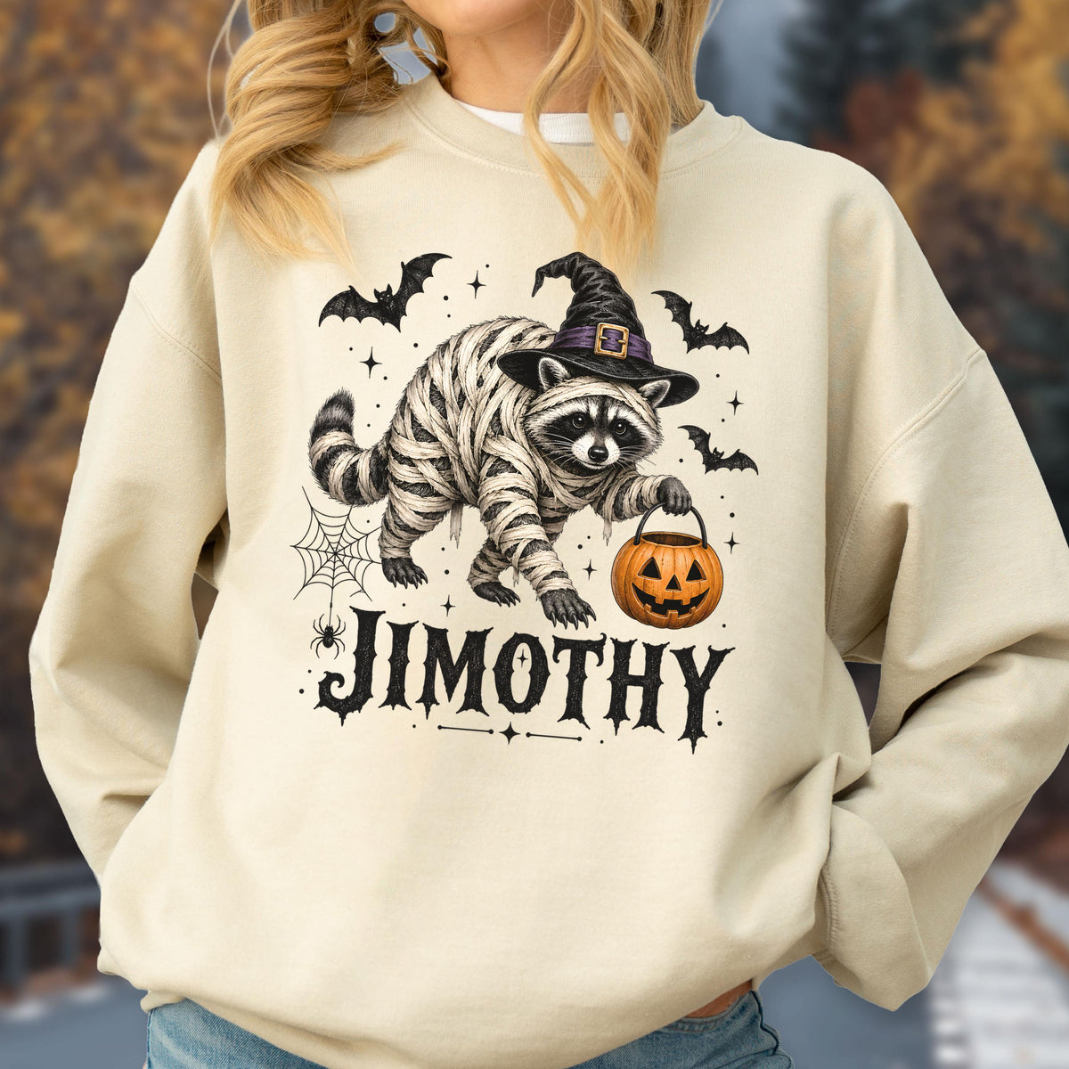 May include: A cream-colored crewneck sweatshirt featuring a graphic of a raccoon wrapped in bandages like a mummy, wearing a black witch hat, and holding an orange jack-o'-lantern bucket. The text JIMOTHY is printed below in black.