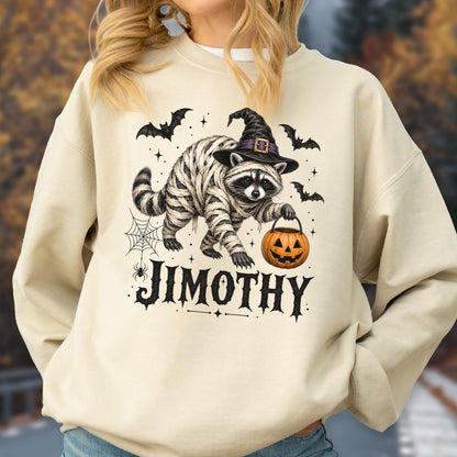May include: A cream-colored crewneck sweatshirt featuring a graphic of a raccoon wrapped in bandages like a mummy, wearing a black witch hat, and holding an orange jack-o'-lantern bucket. The text JIMOTHY is printed below in black.
