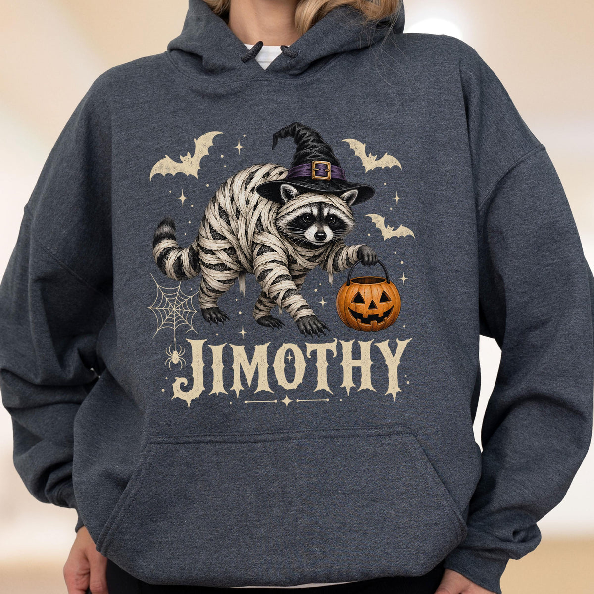 May include: A dark heather gray hooded sweatshirt featuring a graphic of a raccoon wrapped in white mummy bandages, wearing a black witch hat, and holding an orange jack-o'-lantern. The design includes bats, a spider web, and the text JIMOTHY.
