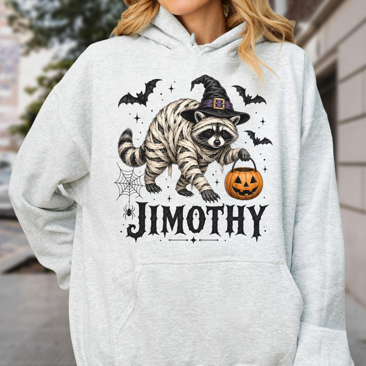 May include: A light gray hooded sweatshirt featuring a graphic of a raccoon wrapped in bandages like a mummy, wearing a black witch hat, and holding an orange jack-o'-lantern bucket. The design includes bats, a spider web, and the name JIMOTHY.