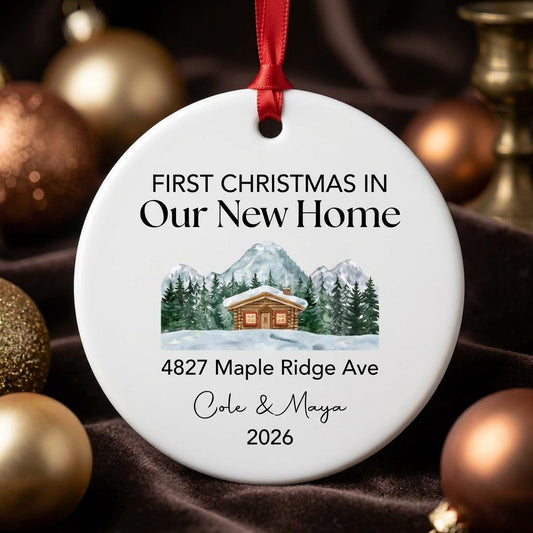 May include: A round white ceramic ornament hanging from a red ribbon. It features the text "FIRST CHRISTMAS IN Our New Home", "4827 Maple Ridge Ave", "Cole & Maya", and "2026" above and below a watercolor illustration of a cabin in snowy mountains.