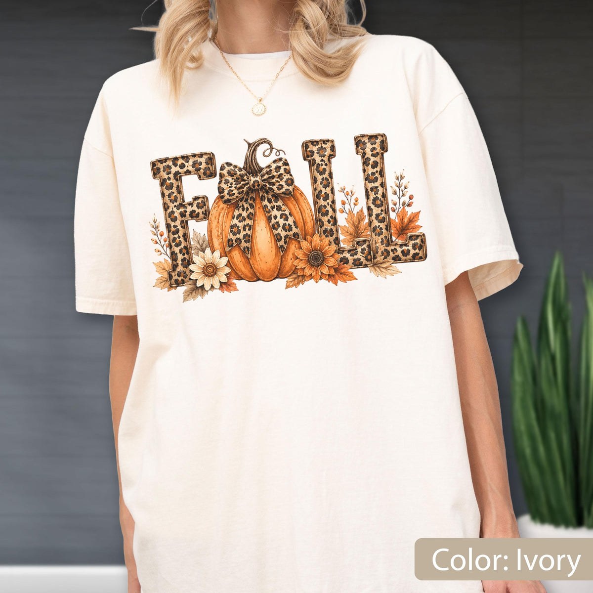 May include: An ivory short-sleeve t-shirt featuring the word FALL in a leopard print pattern. The letter A is replaced by an orange pumpkin with a leopard print bow, surrounded by sunflowers and autumn leaves in shades of brown and orange.