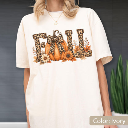 May include: An ivory short-sleeve t-shirt featuring the word FALL in a leopard print pattern. The letter A is replaced by an orange pumpkin with a leopard print bow, surrounded by sunflowers and autumn leaves in shades of brown and orange.