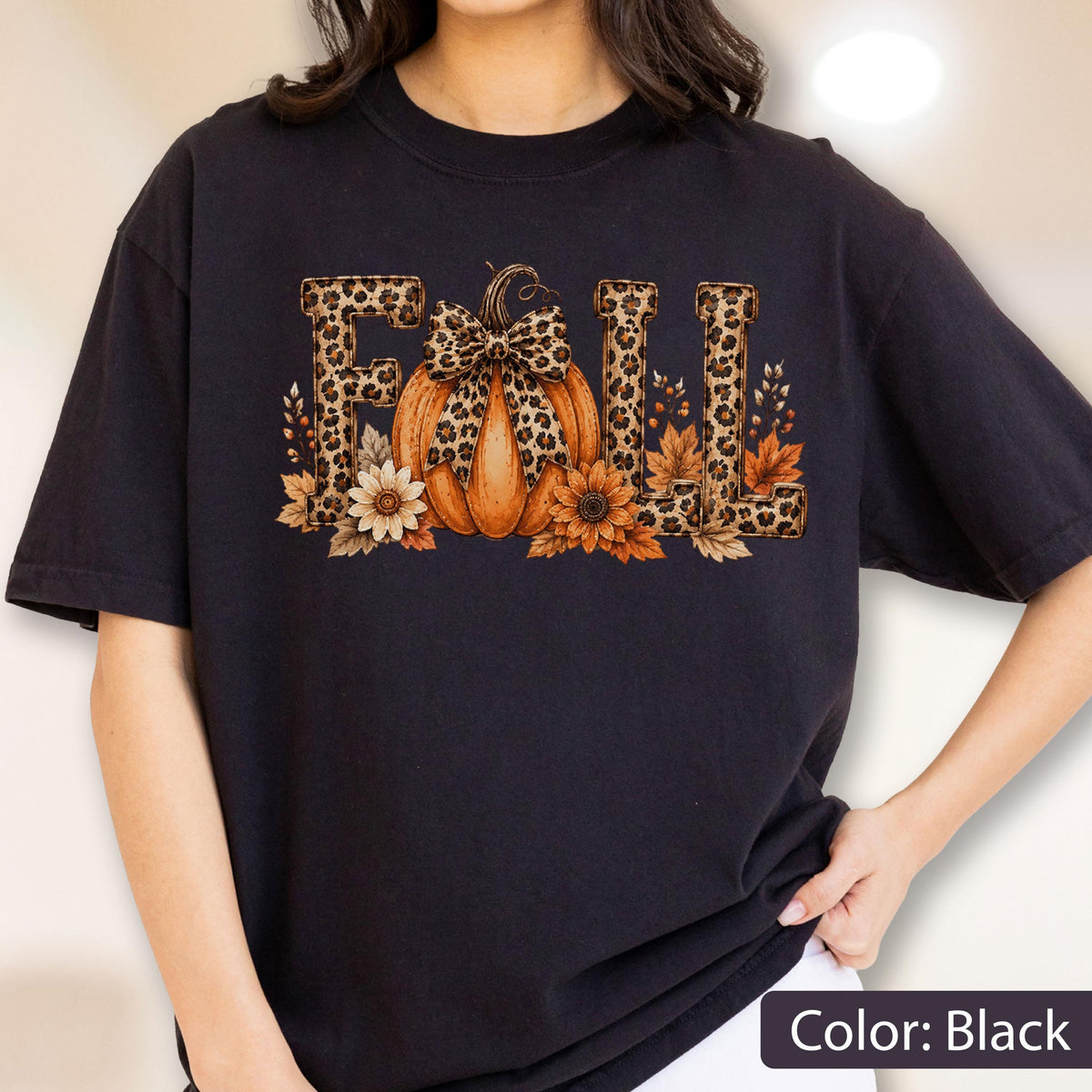 May include: A black t-shirt featuring a graphic print of the word FALL in a leopard print pattern. The letter A is replaced by an orange pumpkin with a leopard print bow, surrounded by orange and brown autumn leaves and sunflowers.