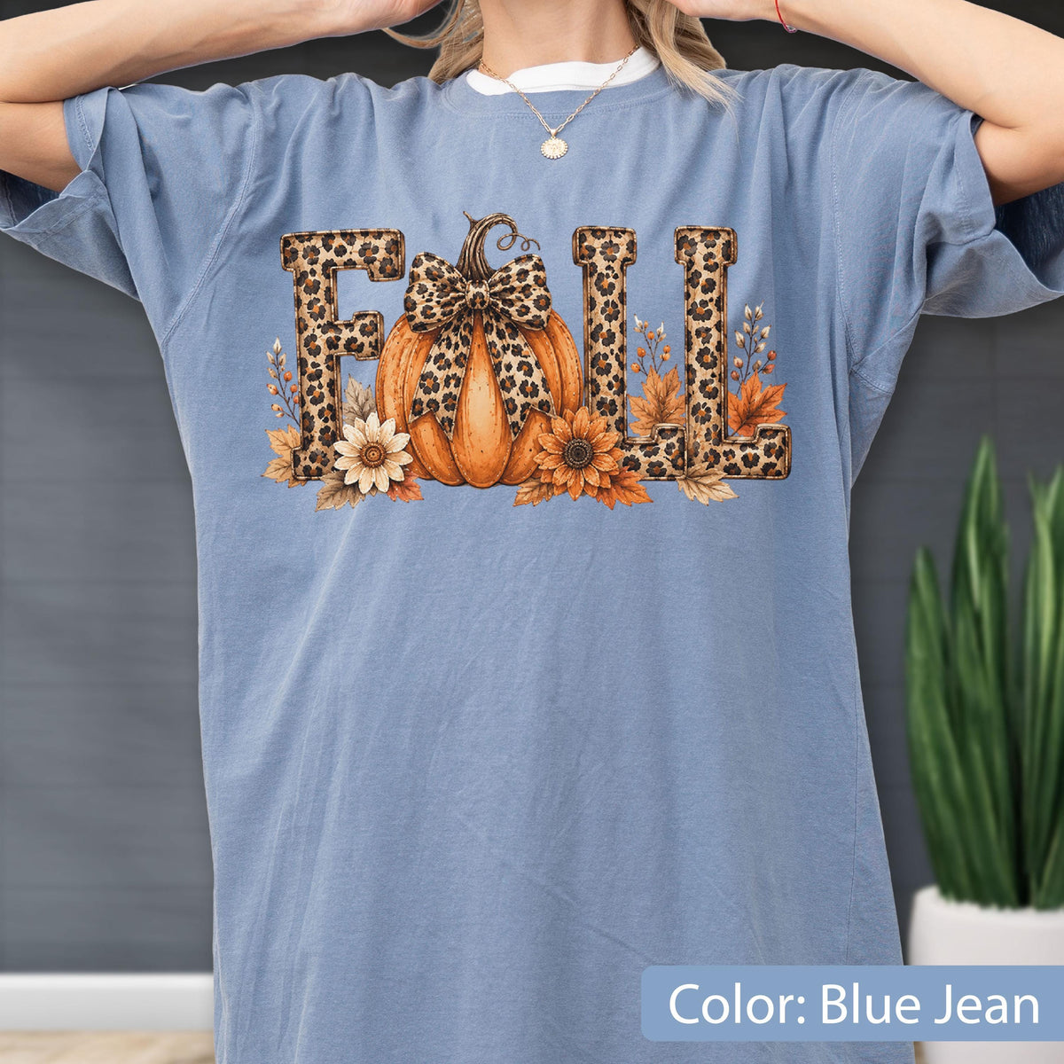 May include: A blue jean colored t-shirt featuring a graphic print of the word FALL. The letters are filled with a leopard print pattern. An orange pumpkin with a leopard print bow sits in the center, surrounded by sunflowers and autumn leaves.