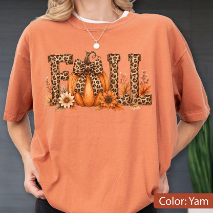 May include: A yam-colored short-sleeve t-shirt featuring a graphic print of the word FALL. The letter A is replaced by an orange pumpkin with a leopard print bow, and the letters F, L, and L feature a leopard print pattern with sunflowers.