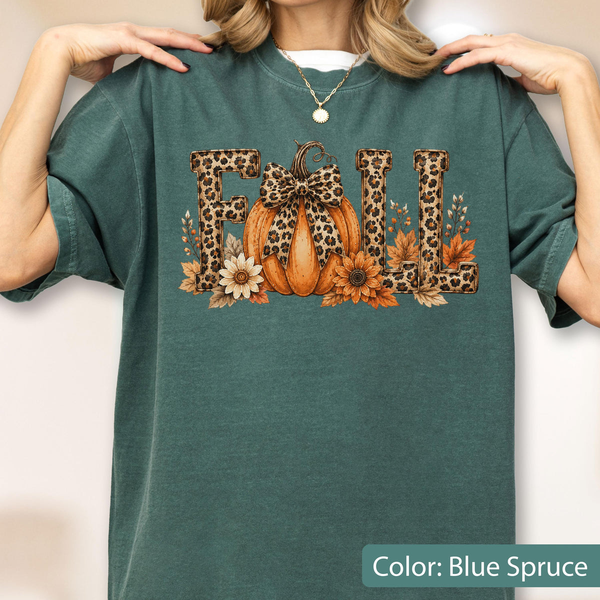 May include: A blue spruce colored t-shirt featuring a graphic print of the word FALL in leopard print letters. An orange pumpkin with a leopard print bow sits in the center, surrounded by orange and white flowers and autumn leaves.