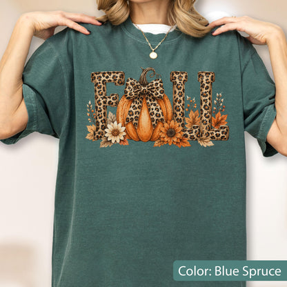 May include: A blue spruce colored t-shirt featuring a graphic print of the word FALL in leopard print letters. An orange pumpkin with a leopard print bow sits in the center, surrounded by orange and white flowers and autumn leaves.