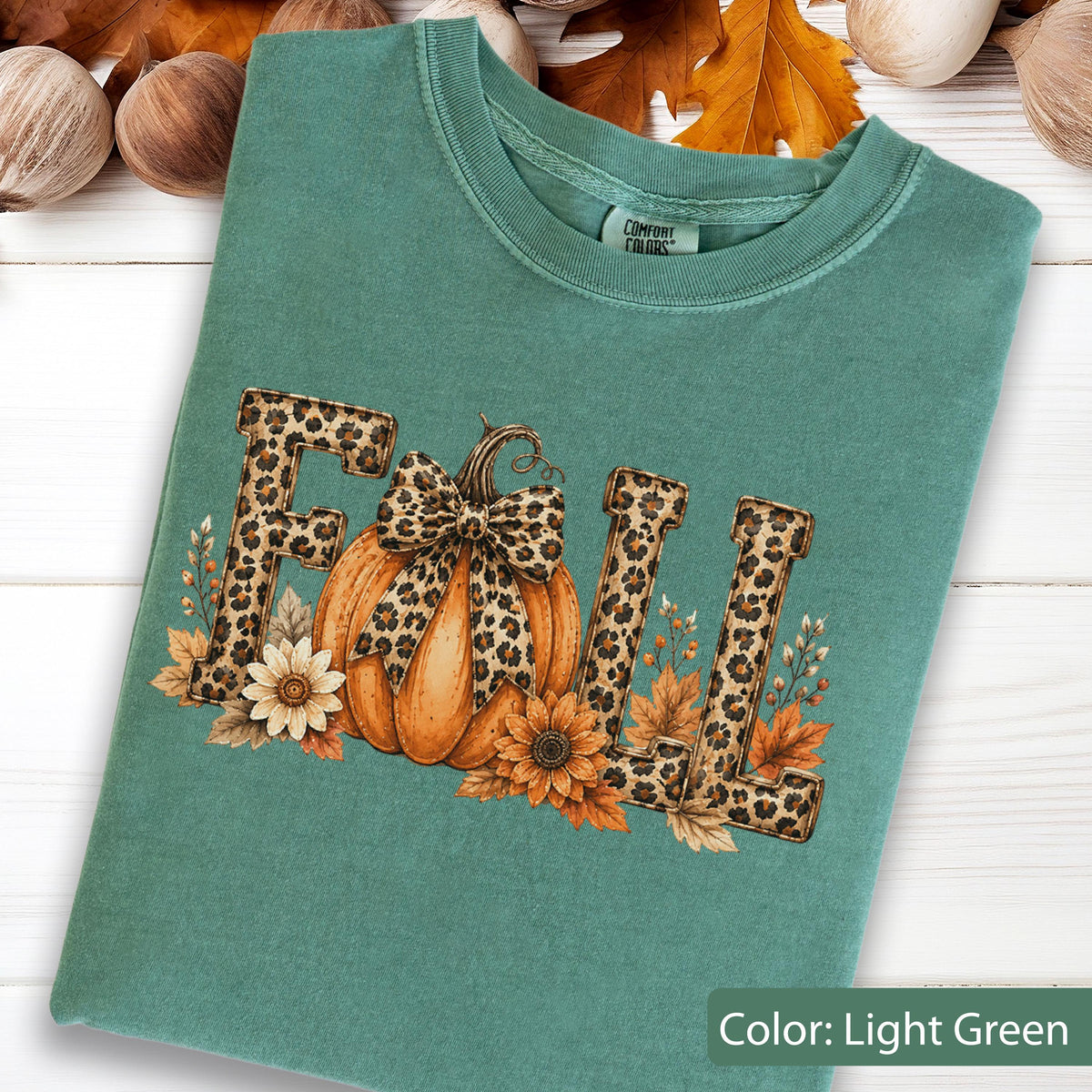 May include: A light green crewneck t-shirt featuring the word FALL in a leopard print pattern. The letter A is replaced by an orange pumpkin with a leopard print bow. The design includes sunflowers and autumn leaves surrounding the text.