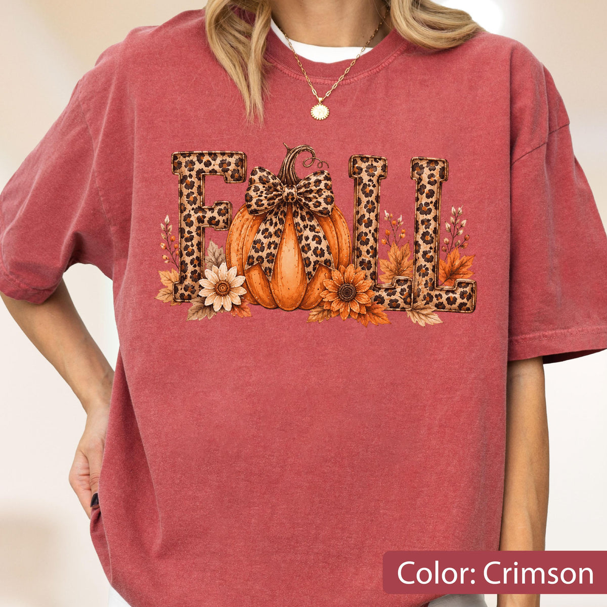 May include: A crimson short-sleeve t-shirt featuring a graphic print of the word FALL. The letters are filled with a leopard print pattern. A central orange pumpkin with a leopard print bow sits between the letters, surrounded by autumn flowers.
